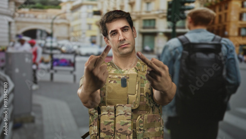 Obraz na plátně Man soldier in camouflage vest and dog tags showing middle fingers on street; defiance protest tension