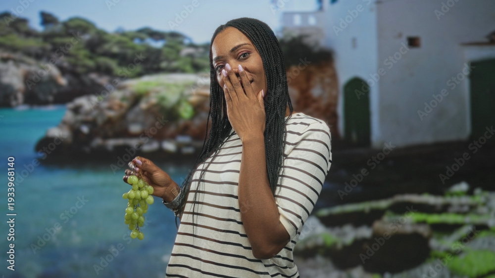 Obraz premium Woman holding a bunch of green grapes while covering her mouth with hand in a studio with seaside backdrop, laughing and smiling candidly; joy.