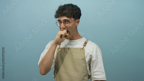 Obraz na płótnie Young hispanic man wearing apron and glasses biting his fist in studio against pale blue wall with soft lighting; anxiety uncertainty