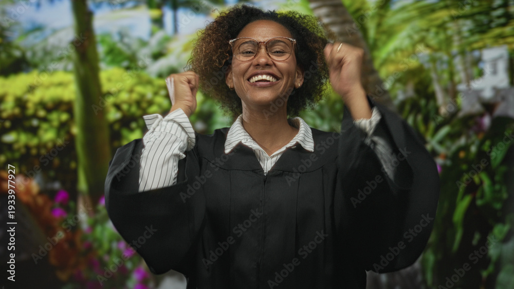 Fototapeta premium Woman in black robe with fists raised smiling broadly and wearing glasses in forest with lush green plants; academic achievement celebration.