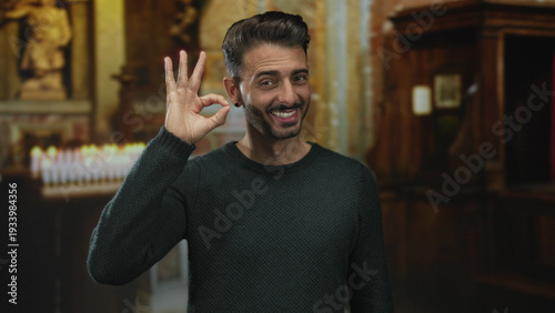 фотографія Young smiling man shows ok sign with hand in candlelit ancient building corridor indoors; approval