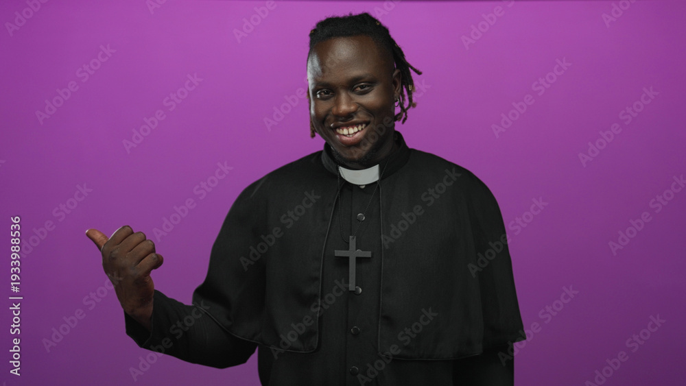 Obraz premium Young african man priest pointing thumb to side wearing cross necklace in purple studio; welcoming joy community.