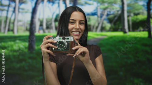 Fotografi Woman smiling with vintage camera in park under sunlit trees showcasing blissful outdoor photography moment