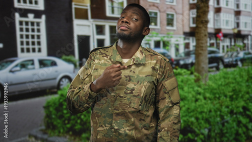 Fotografía Man in camouflage soldier uniform points to chest with hand on street in front of building and parked cars; pride duty