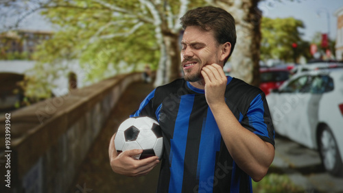 Billede på lærred Man holding soccer ball and touching jaw for toothache on street while squinting in team jersey; injury frustration