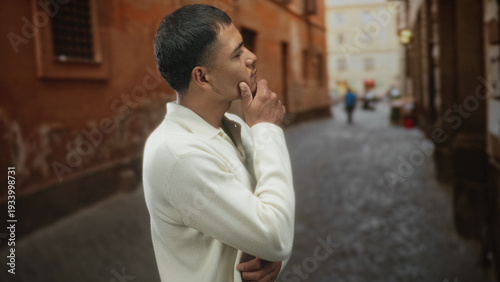 Εκτύπωση καμβά Man touching chin with hand in thinking pose on a narrow cobblestone street, wearing white sweater and looking sideways; quiet contemplation