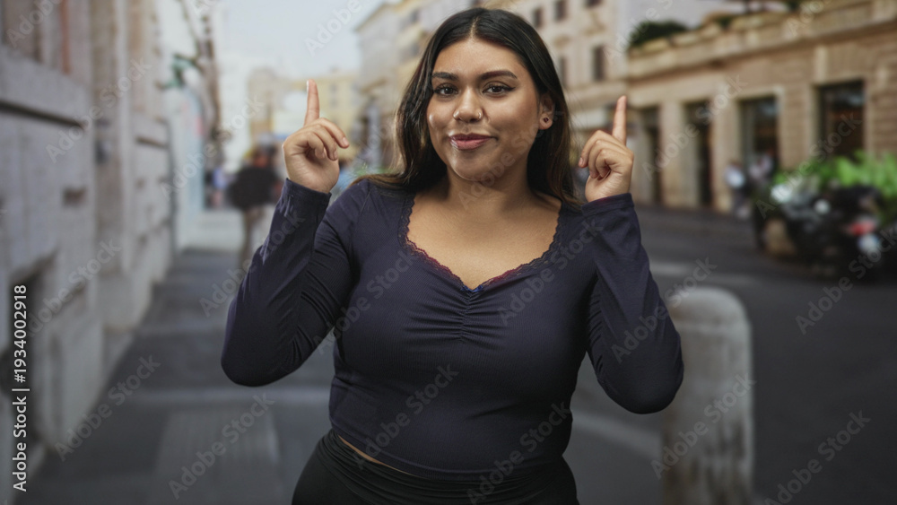 Fototapeta premium Plus size woman points index fingers upward on a city street, wearing navy top with lace neckline, smiling with closed eyes; joy confidence.