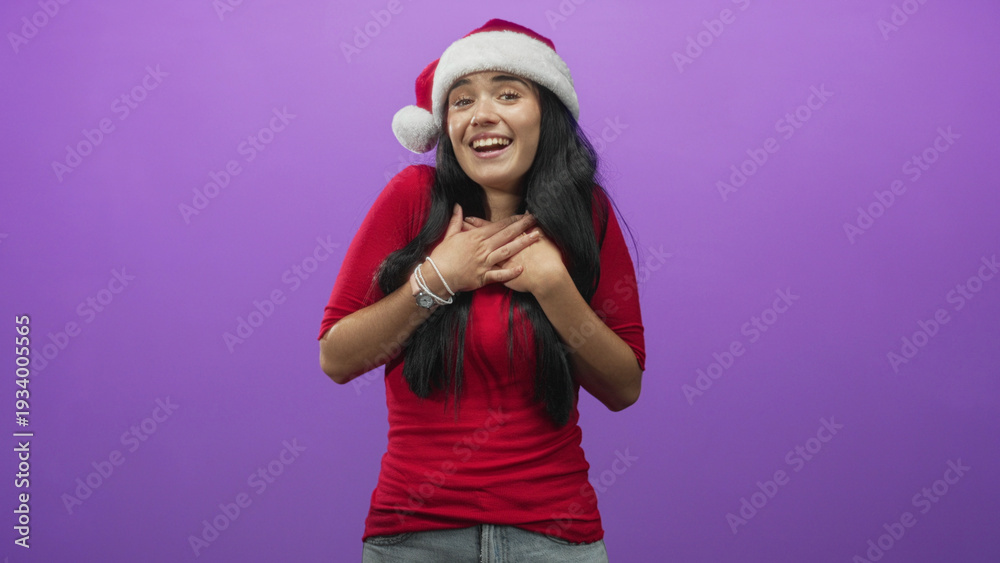 Fototapeta premium Young hispanic woman wearing santa hat and red shirt clasps hands to chest while smiling with closed eyes against a purple backdrop in studio; festive joy.