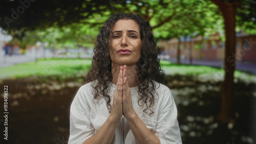 Fotografía Woman with praying hands gesture in a leafy sunlit park with blurred building beyond; prayer faith serenity