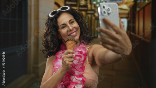 Quadro su tela Middle aged hispanic woman wearing pink bikini and hawaiian lei holding ice cream cone and smartphone for selfie in narrow street arcade; joy vacation leisure