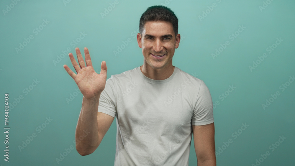 Naklejka premium Man smiling and waving hand to camera in teal studio wearing casual white t shirt with direct eye contact; friendly greeting.