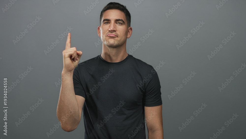 Fototapeta premium Man wearing black tshirt pointing index finger upward in gray studio; determination resolve focus intent.