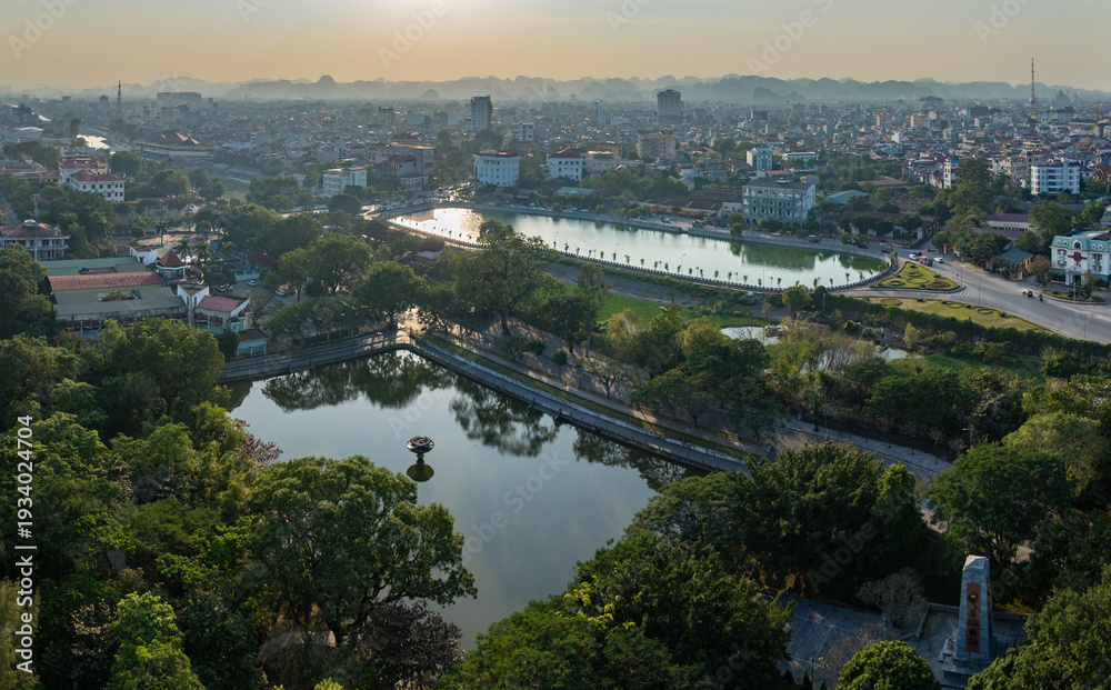 Fototapeta premium Panoramic aerial view of Park Thuy Son, Ninh Binh City, Vietnam, featuring its serene lakes, urban architecture, and the historic War Memorial at sunrise.