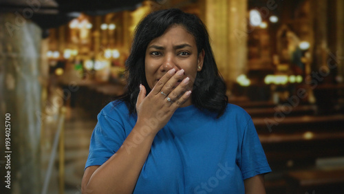 Bild auf Leinwand African american woman covers mouth with hand gesture next to wooden pew inside church interior; surprise