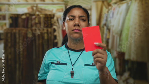 Canvas-taulu Referee woman holds up bright red card among hanging clothes in indoor retail shop while looking at camera; warning