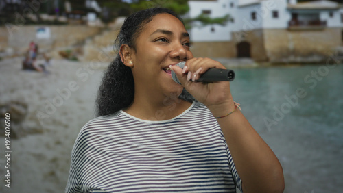 Konstfotografi Woman singing into handheld microphone by seaside building with a bright smile and closed eyes; joyful celebration