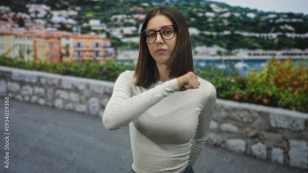 Fototapeta premium Woman in white long sleeve shirt wearing glasses points thumb to chest on a seaside street by a stone wall and marina; confidence assertiveness contemplation.