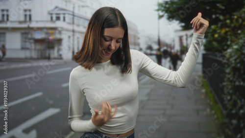 Foto Young woman in white top and jeans dancing with raised arm and visible bellybutton on a street sidewalk near a white building and hedge; carefree movement
