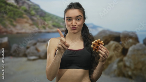 Foto Young hispanic woman holding waffle points finger upward and puckering lips on sunny beach; rejection