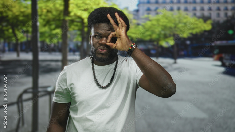Naklejka premium Man in white tshirt making ok sign with hand over eye on street, wearing beaded necklace and bracelets, facing camera with slight smile; playful curiosity.