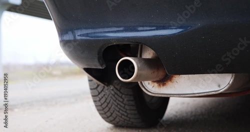 Close-up view of a car exhaust pipe showing gradual angle change, revealing tire tread and rust details, set against an outdoor backdrop with a blurred structure