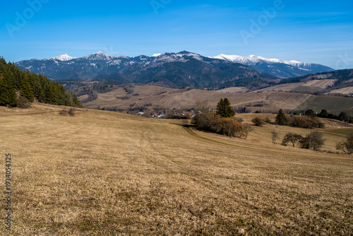 Spring landscape of meadows in the foothills and a village in the valley and snow-capped peaks in the background.