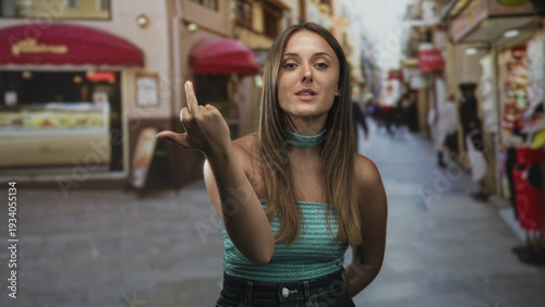 Woman shows middle finger toward camera on a narrow pedestrian street with shop awnings and blurred passersby; defiance.