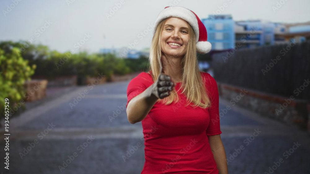 Fototapeta premium Blonde woman extends right hand for handshake on street in front of residential buildings, wearing santa hat and red top; friendly holiday greeting.