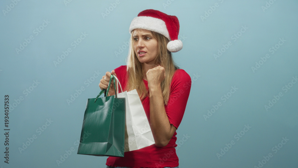 Obraz premium Blonde woman wearing santa hat and red top holds green and white shopping bags and touches chin in studio; holiday uncertainty.