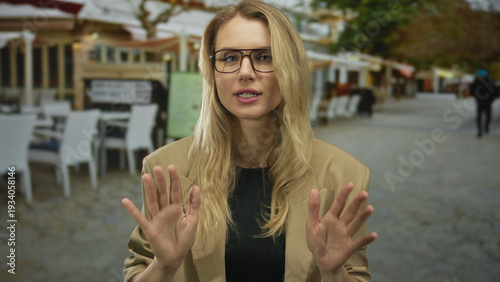 Quadro su tela Woman gesturing calmly on a city street terrace in an outdoor restaurant setting wearing glasses and a beige blazer with blonde hair