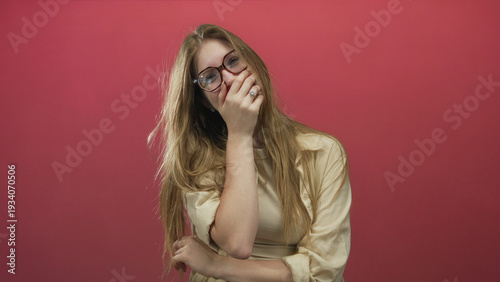 Obraz na plátně Young blonde woman with long hair and glasses covers mouth with hand while tilting head in studio; amusement