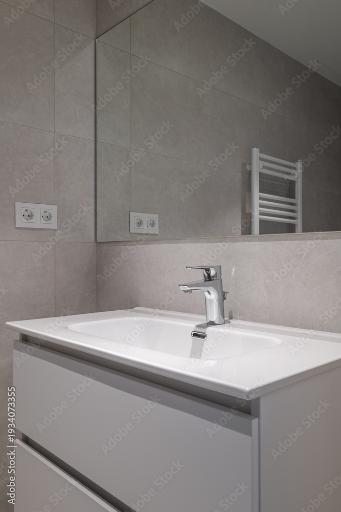 Fototapeta premium Modern bathroom interior with a sleek white sink, chrome faucet, and gray tiled walls. A mirror reflects the minimalist design and a towel radiator is visible.