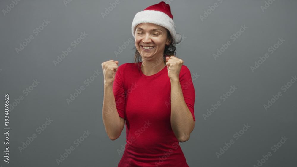 Fototapeta premium Woman in red shirt and santa hat clenches fists, raises arms and smiles in studio; holiday excitement.