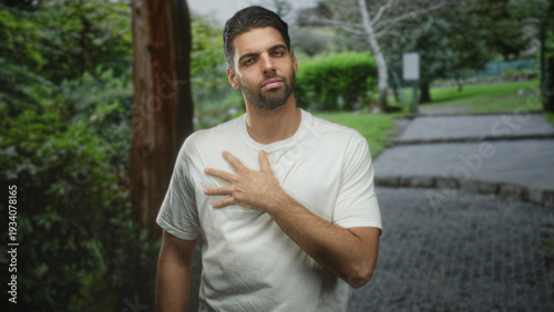 Hispanic man with beard presses hand to chest on forest path with closed eyes and pensive expression; quiet contemplation.