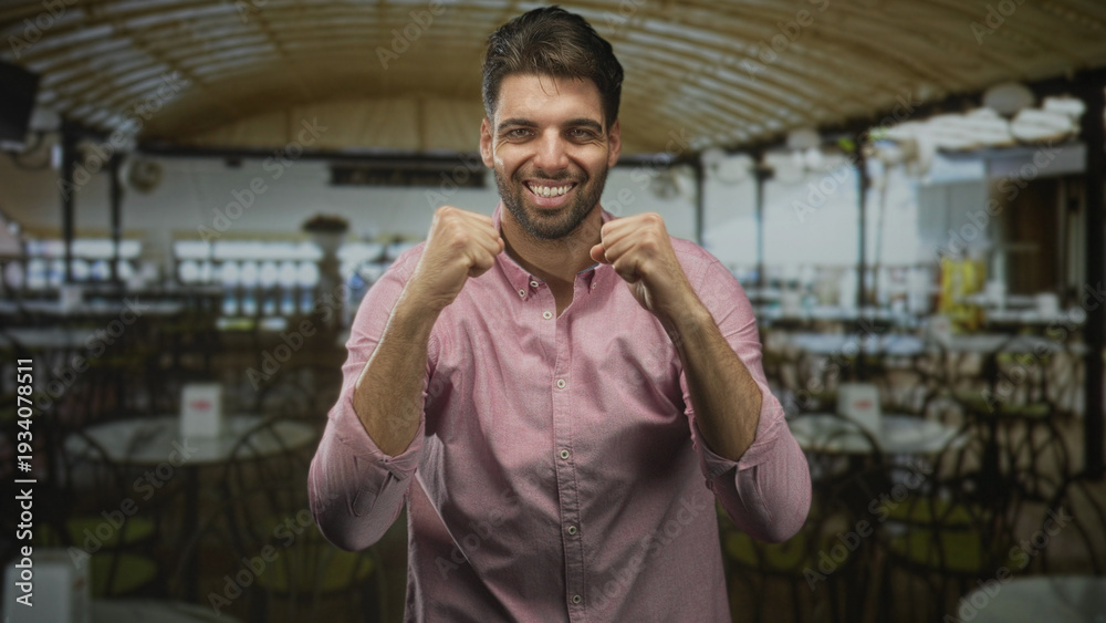 Fototapeta premium Hispanic man with beard raises clenched fists and smiles amid empty cafe tables under arched ceiling in a building; joy celebration.