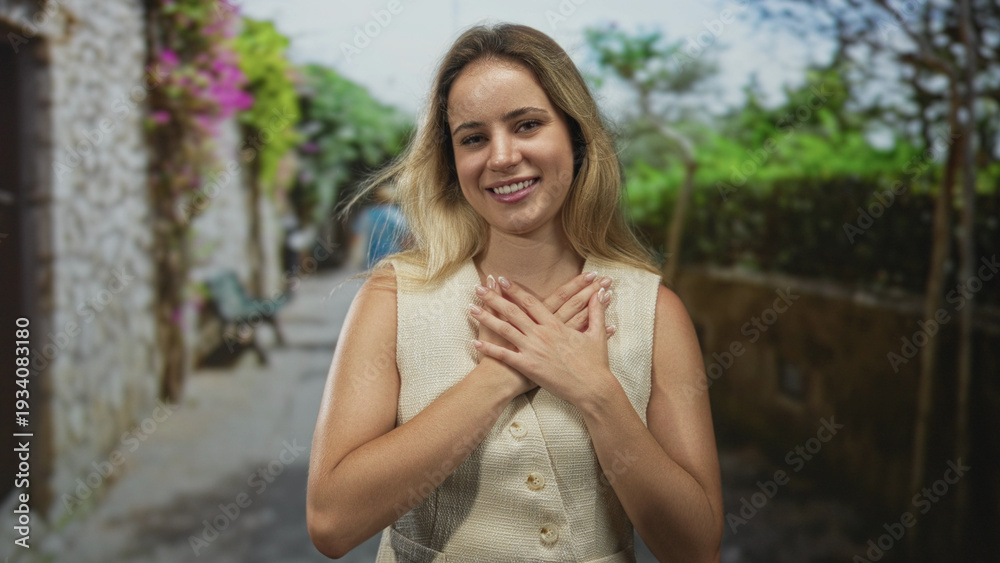 Naklejka premium Woman blonde smiling with hands crossed on chest on a street with stone wall and greenery; gratitude connection.