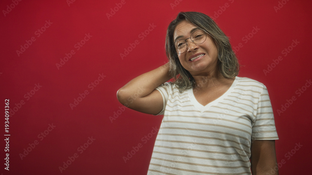 Fototapeta premium Woman touches neck with hand in red studio, wearing striped t shirt and glasses showing a wry grimace; mild embarrassment.