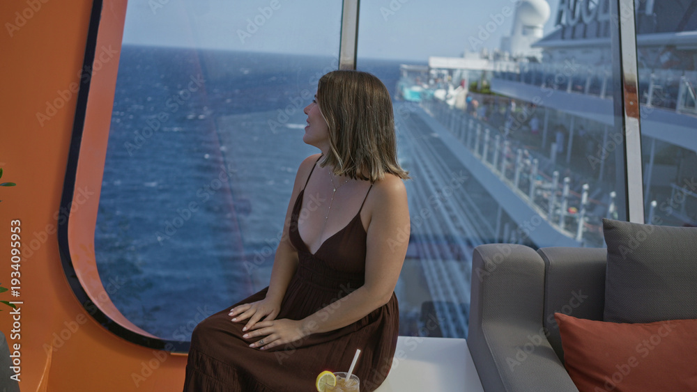Fototapeta premium Woman brunette young hispanic sitting with hands on lap and visible cleavage wearing brown dress by cruise terrace window with ocean view; serenity relaxation.