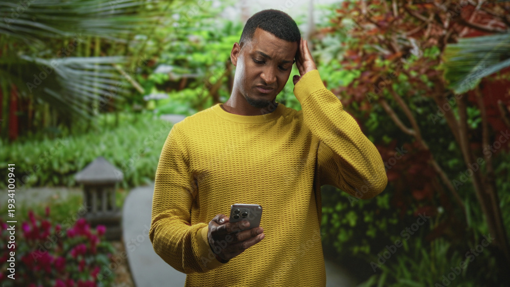 Fototapeta premium Black man in yellow sweater holds smartphone, frowns and rubs his temple while checking messages on a street garden path; confusion tech problem.