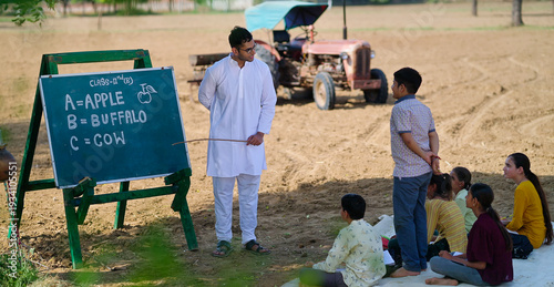 Indian young adult gen z male man guy talk speak teach to villager child day under tree rural area. Happy girl boy group sitting on mat do study look board learn read English at open class field place