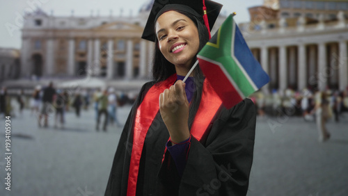 Photos Young woman waves south african flag while wearing black graduation gown and cap in front of vatican building; pride accomplishment celebration