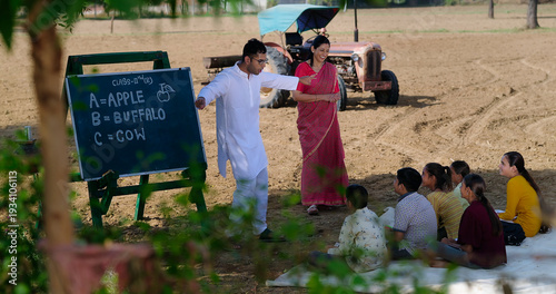 Indian happy young adult male man do dance teach to girl boy standing under tree rural ground area group sitting on mat look woman staff tutor learn enjoy new day have fun joy open class field place