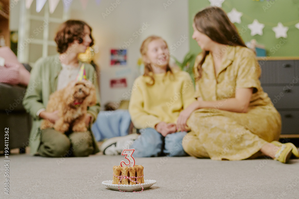 Obraz premium Caucasian child sitting between two Caucasian women on floor, smiling and holding hands, small dog with party hat sitting nearby, birthday cake with number three candle in foreground