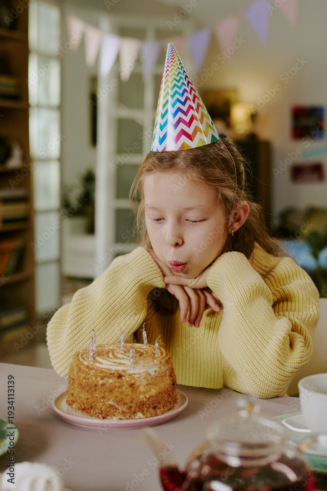Fototapeta premium Vertical of Caucasian girl child wearing party hat blowing out candles on birthday cake while sitting at table indoors, hands resting under chin, celebrating birthday in home setting