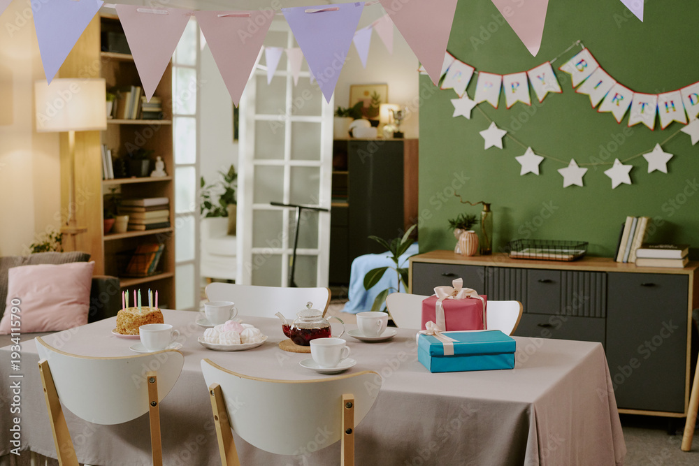 Obraz premium Birthday party table setting featuring cake with candles, wrapped gifts, teacups, and pastries arranged on table, pastel bunting and happy birthday banner decorating background wall