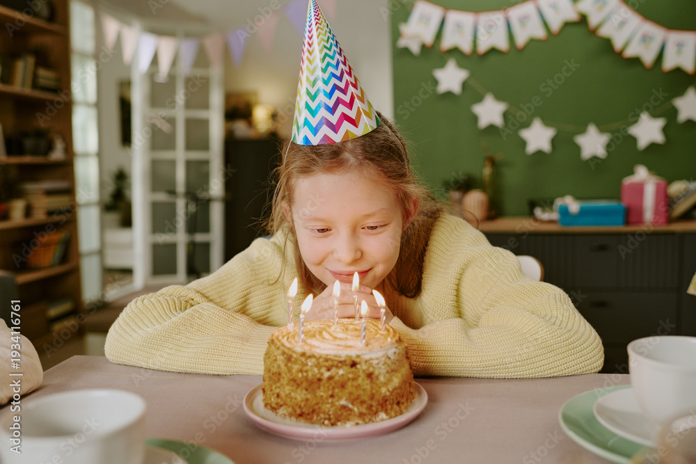Obraz premium Portrait of Caucasian girl child wearing party hat leaning forward blowing out candles on birthday cake during celebration at home, smiling and appearing joyful with cake in foreground