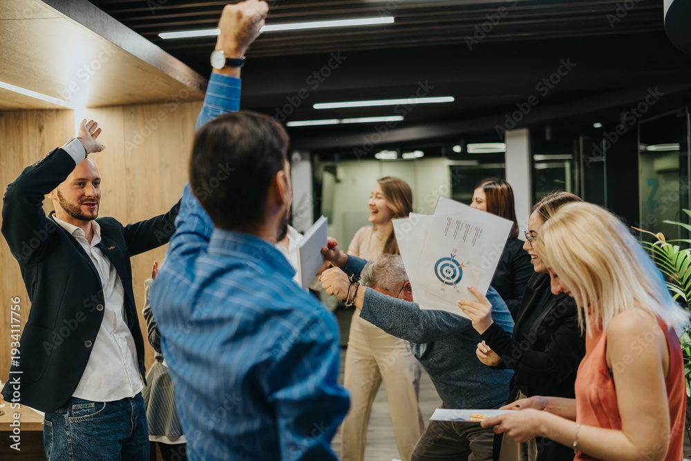 Obraz premium Happy professionals celebrate in an office, raising arms and sharing documents. A diverse team cheers together in a modern workspace during a project milestone.