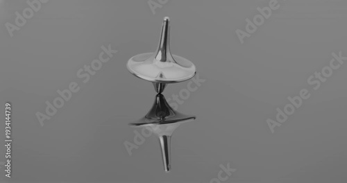 Metallic totem in initial steady rotation, gradually increasing wobble and tilt toward collapse against a mirrored backdrop.