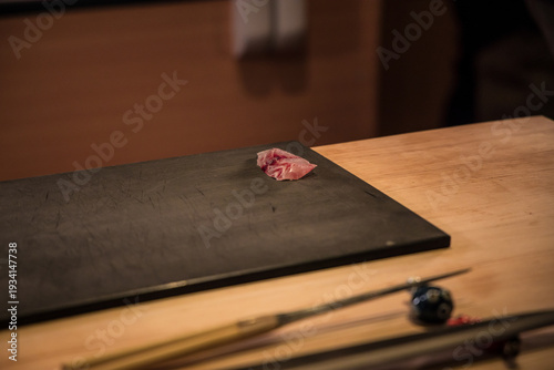 A single slice of fresh sashimi rests on a dark board at a wooden counter. Soft focus tools lie nearby, framing a quiet preparation moment at a sushi bar.