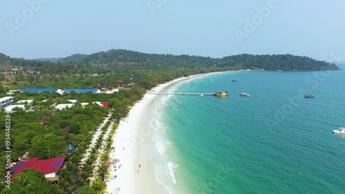Scenic aerial view of Long Set Beach with turquoise water, white sand, and lush greenery. Boats are anchored near a wooden pier extending into the sea.